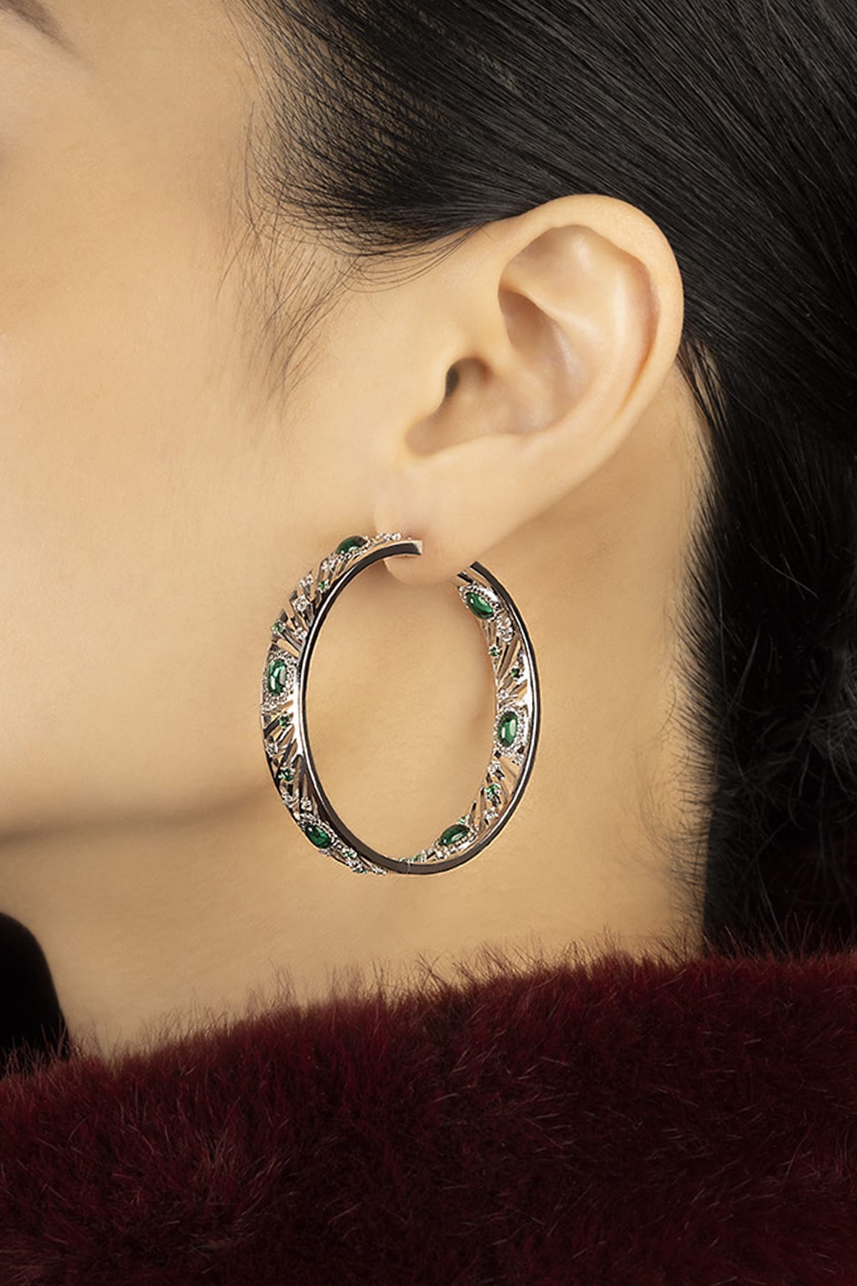 Woman wearing APM hoop earrings, emerald green stones glowing with bold elegance.