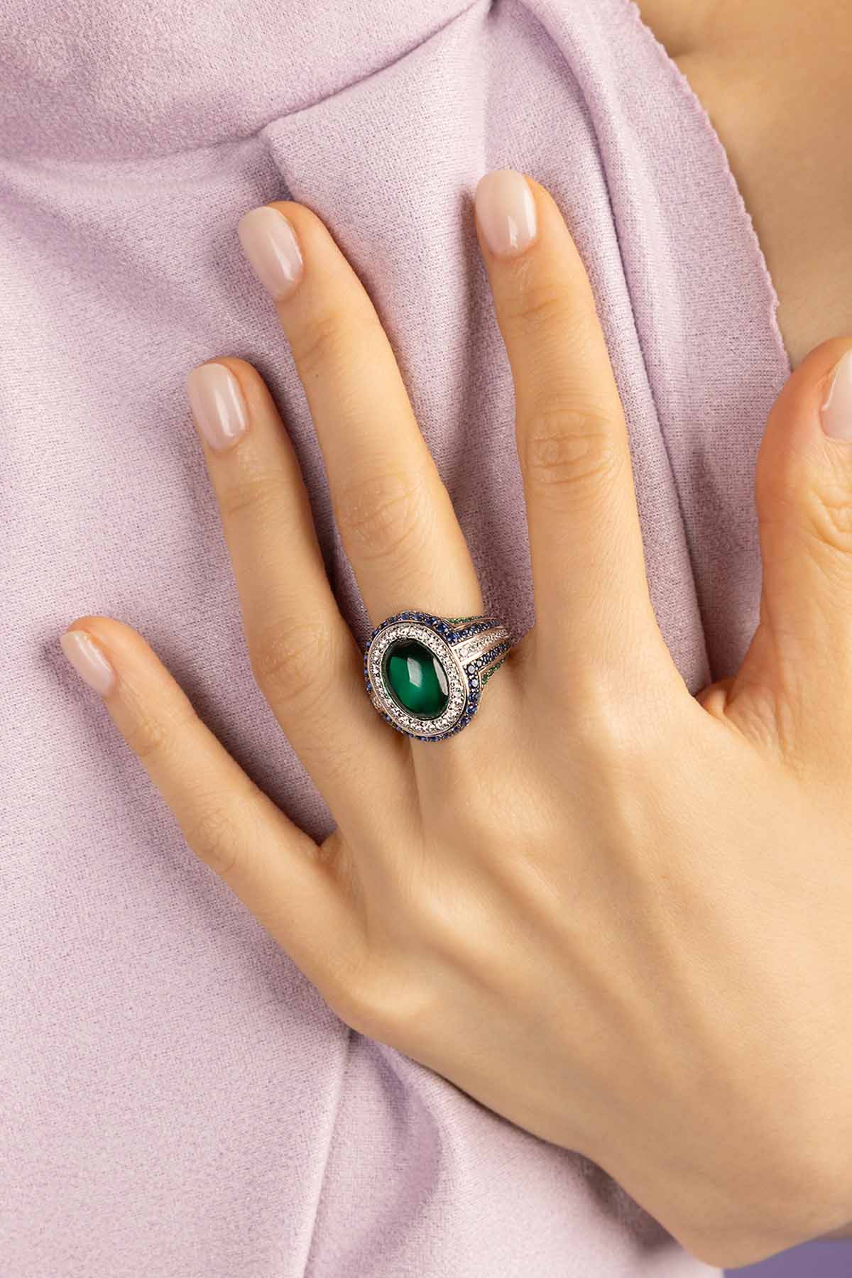 Hand wearing a ring with a green gemstone on a light purple fabric background