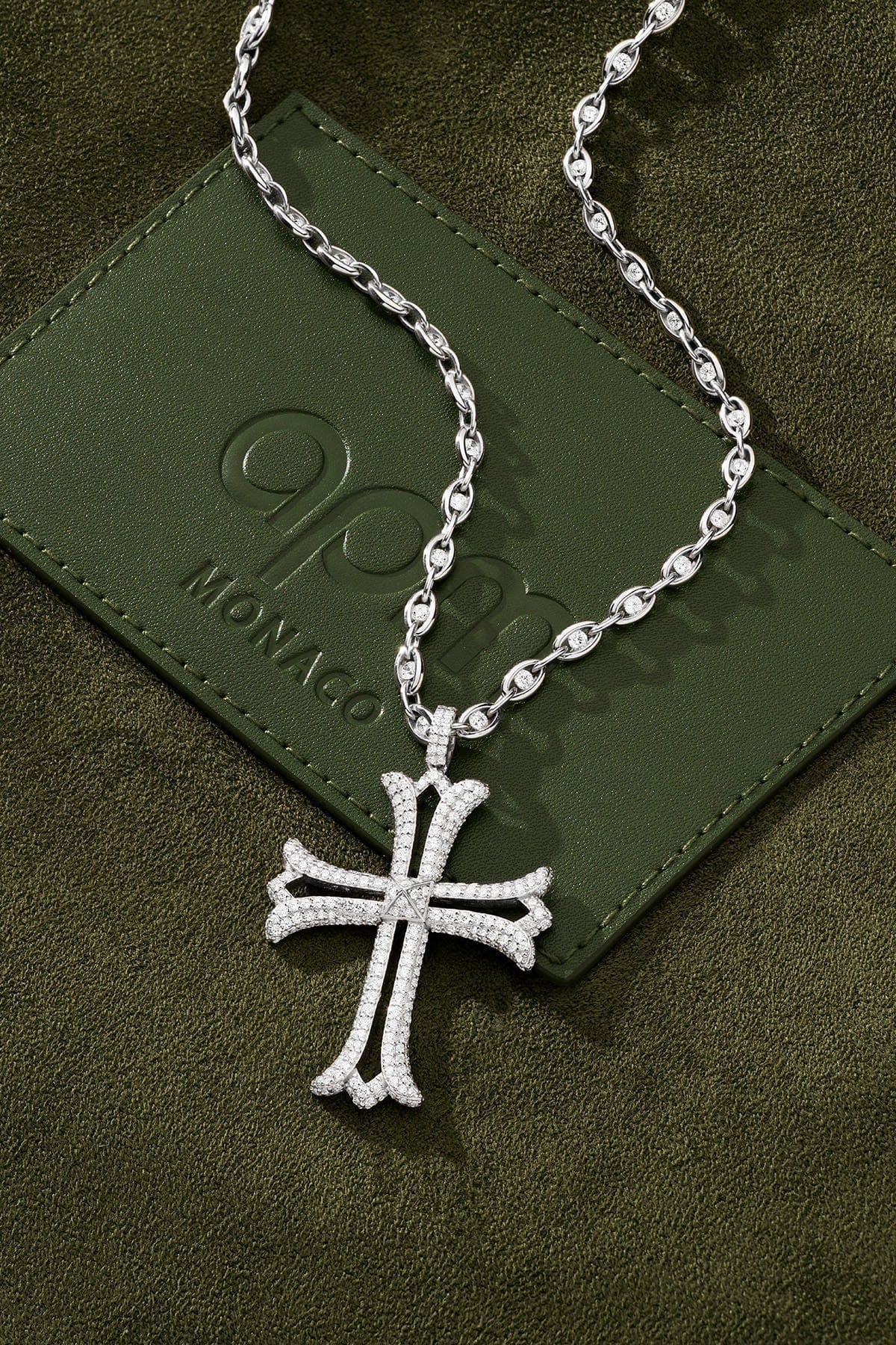 Silver cross necklace on a green textured surface with a brand logo.