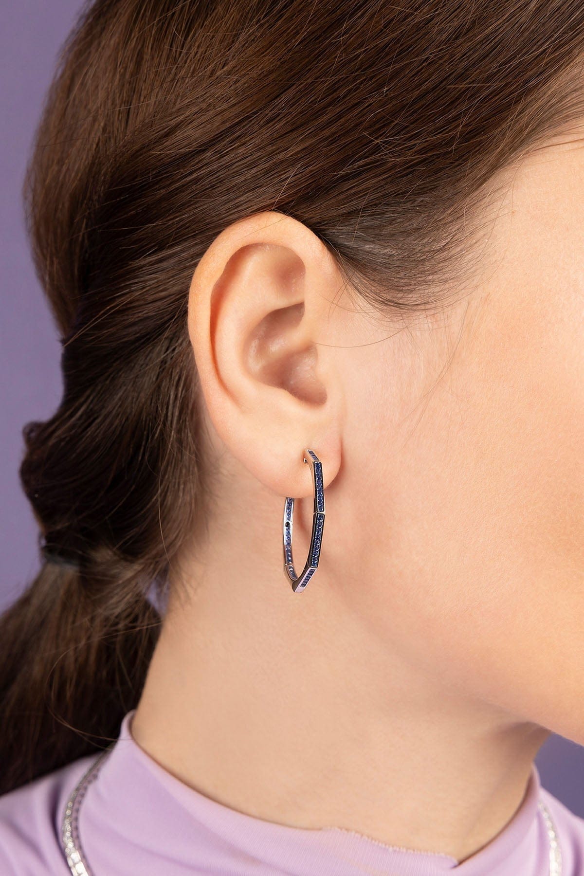 Close-up of an ear wearing a silver hoop earring with a purple background