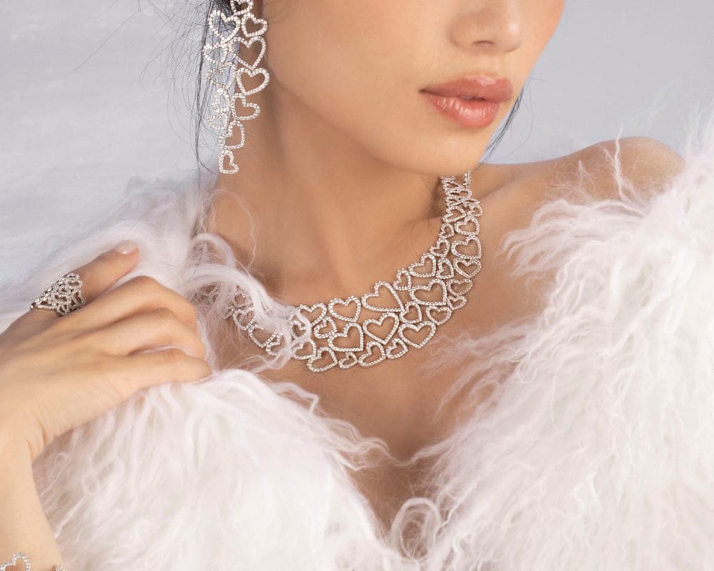 Close-up of a woman wearing APM MONACO silver heart-shaped necklace and earrings with a white feathered garment.