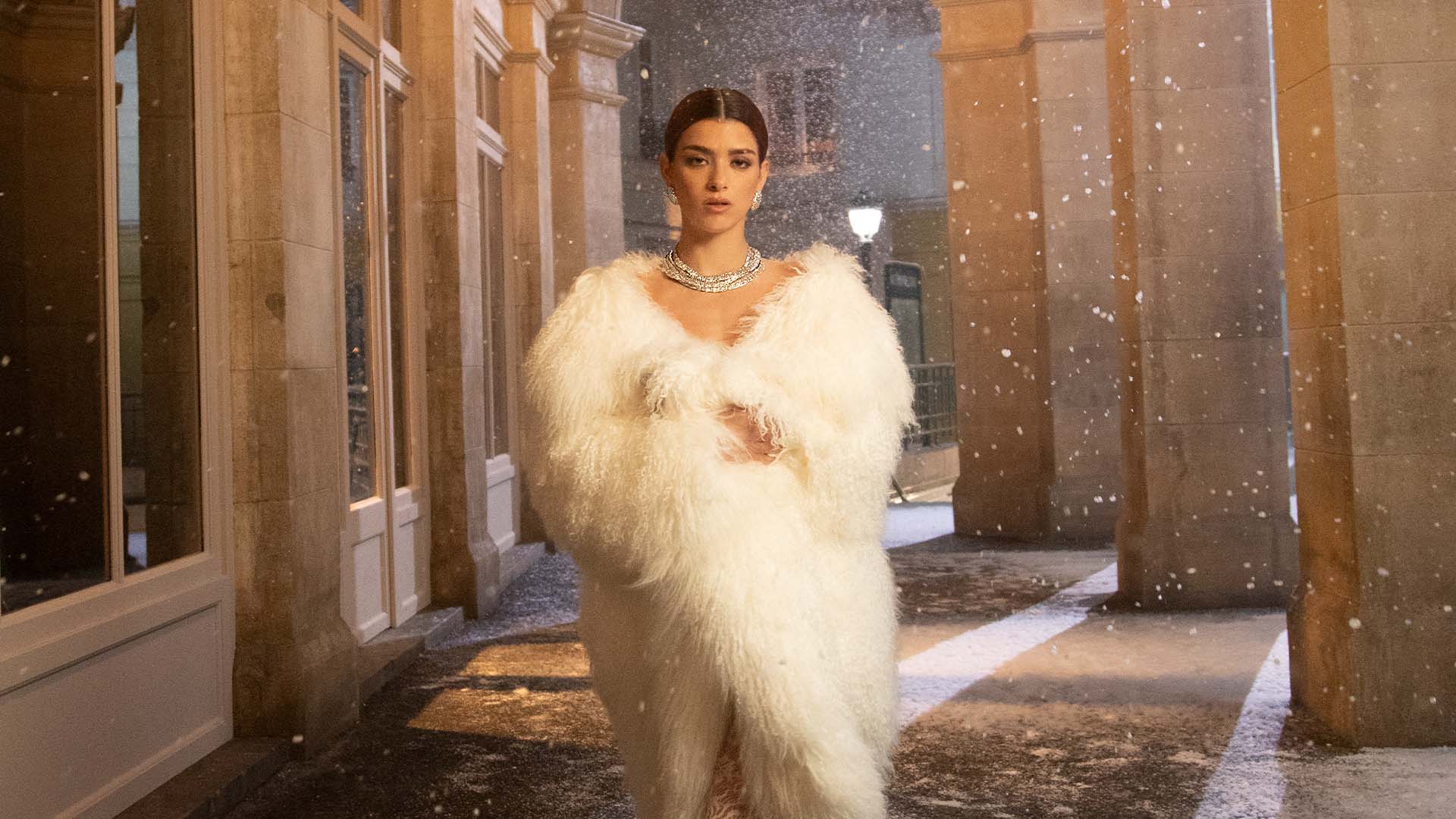 Woman in a white feathered outfit with APM MONACO Ice jewelry set standing in a snowy street with classical architecture.