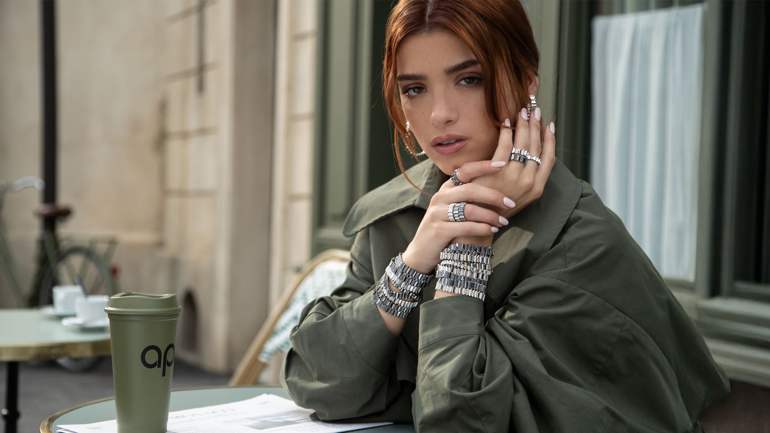 Woman sitting at an outdoor cafe table with a green cup with APM MONACO logo, wearing multiple APM MONACO up and down rings and bracelets.