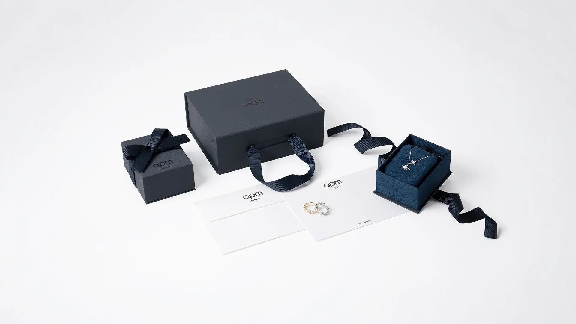 A jewelry gift set box, featuring a navy blue presentation box, a smaller matching box with a ribbon, an open box displaying a silver necklace, a pair of silver rings, and branded white cards on a clean white background.