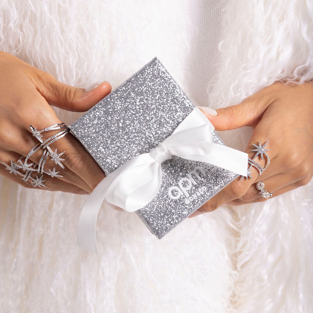 APM Monaco Silver glittery box with a white bow held by hands wearing star-shaped rings on a soft white background