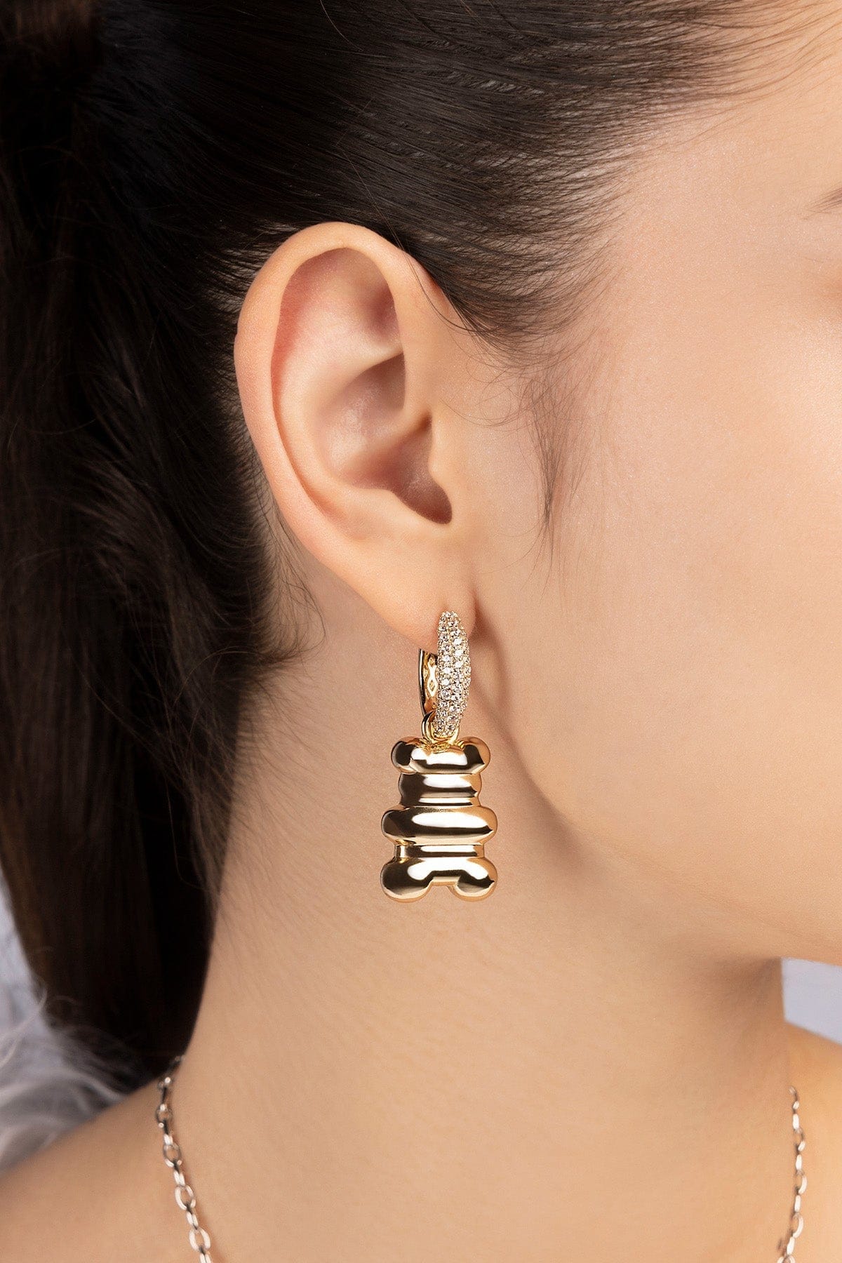 Close-up of a person wearing yellow gold hoop earrings with xl yummy bear design from APM MONACO against a neutral background