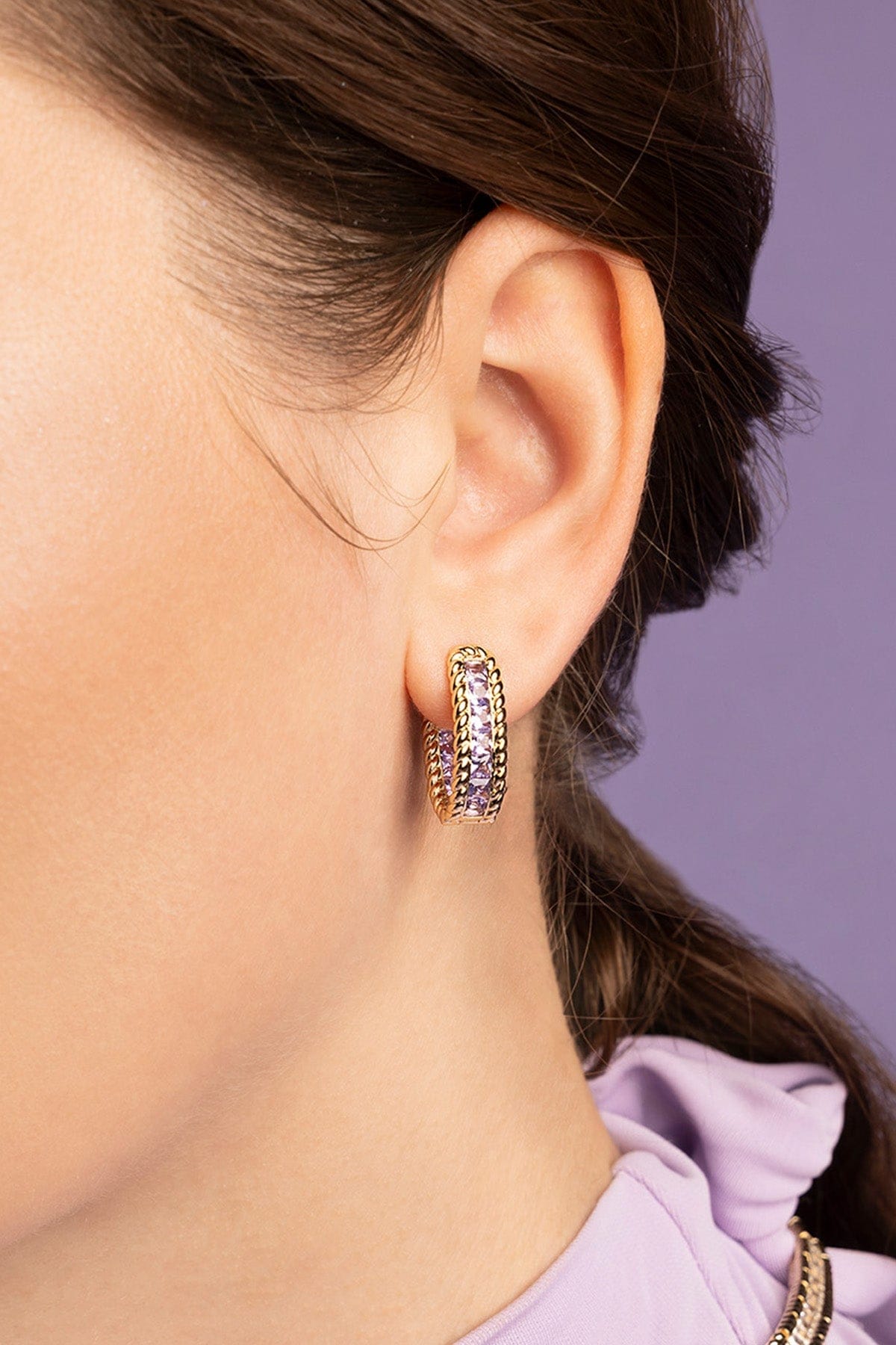 Close-up of an ear wearing a gold hoop earring with purple stones against a blurred background
