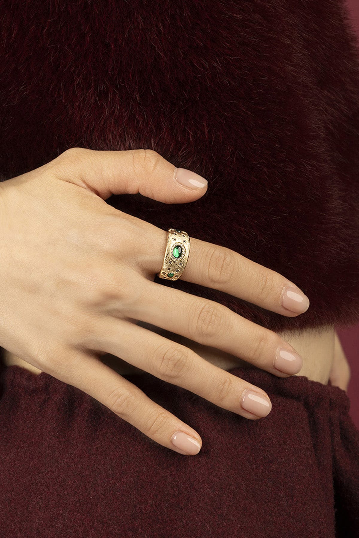 Woman wearing APM Ring, a golden statement with emerald allure against burgundy warmth.