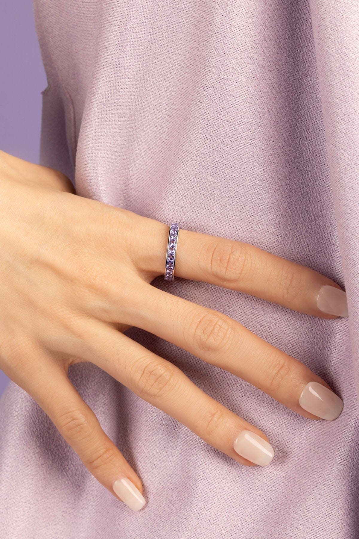 Hand wearing a ring with purple zirconia stones against a light purple background