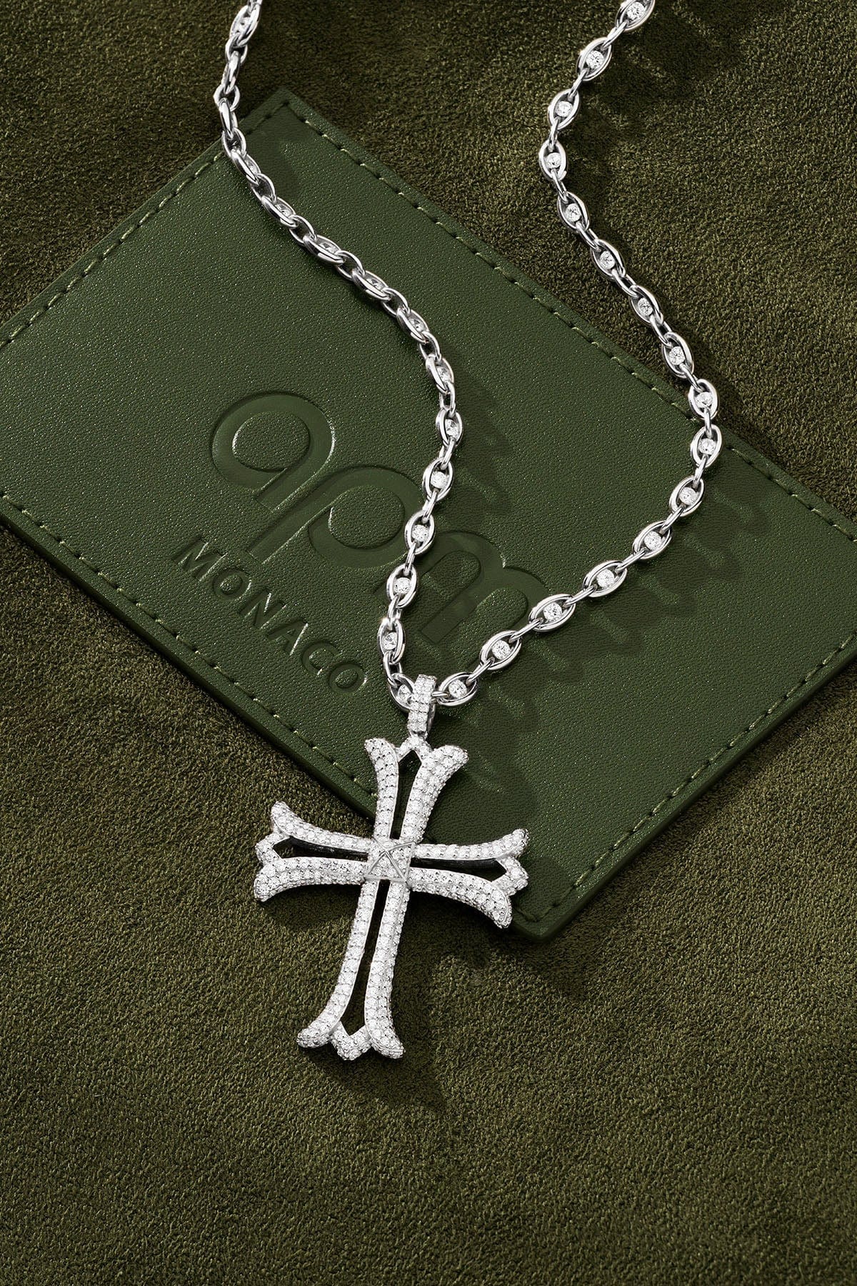 Silver cross necklace on a green textured surface with a brand logo.