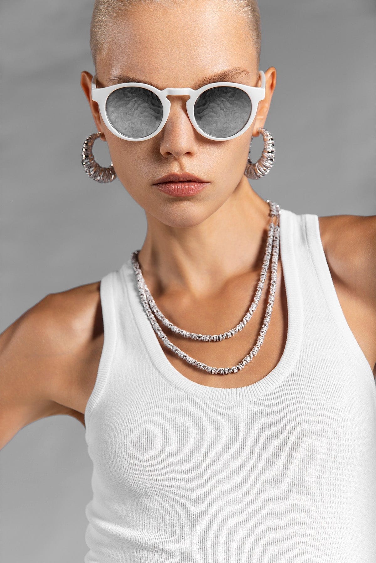 Person wearing white sunglasses and layered APM MONACO silver necklaces on a gray background