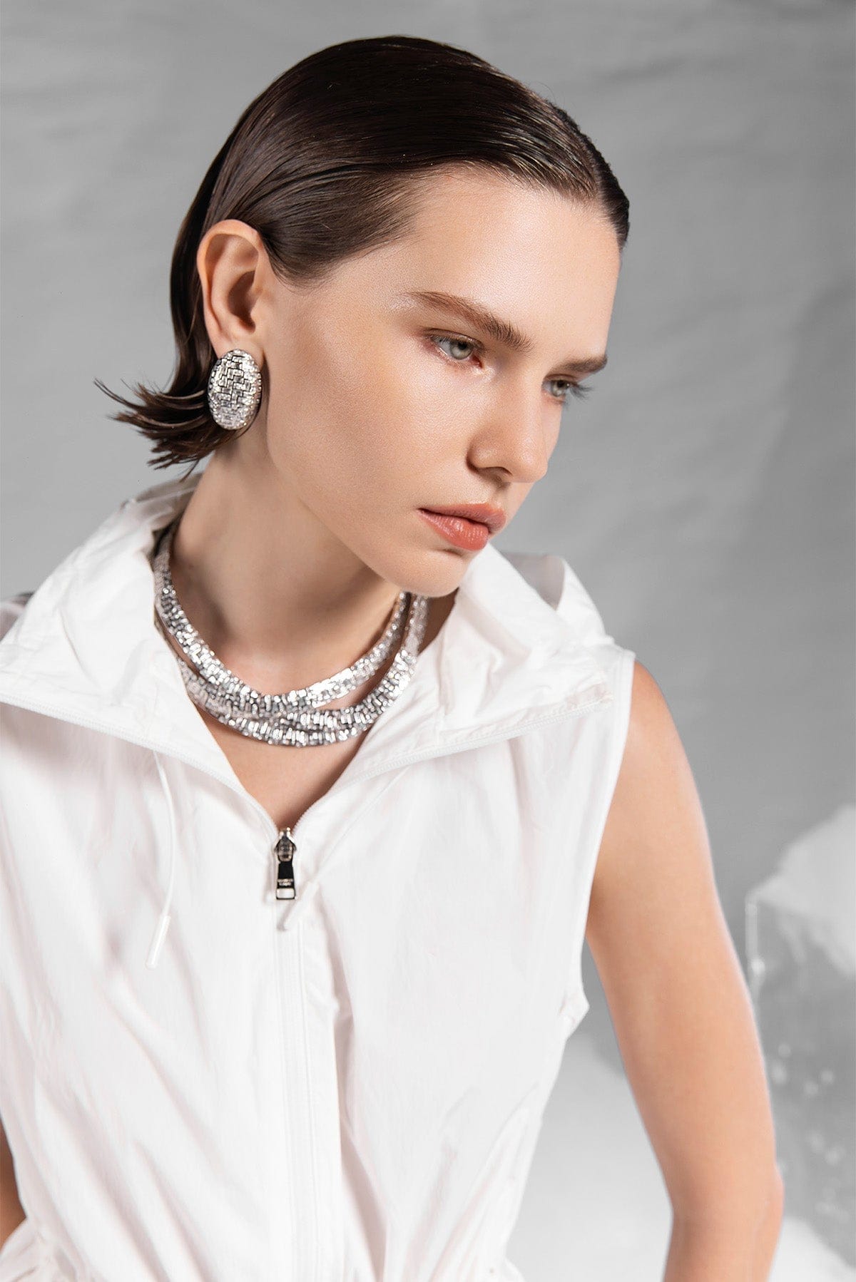 Woman wearing a white outfit with a sparkling APM MONACO necklace and earrings against a gray background