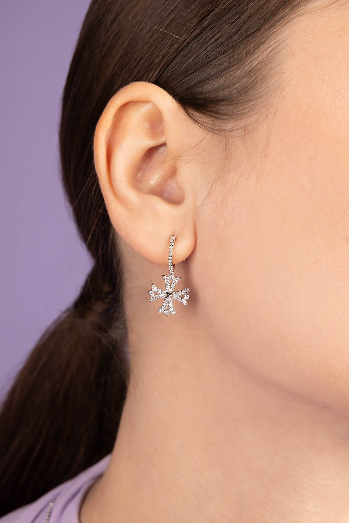 Close-up of a person wearing APM MONACO silver Cross Asymmetric Hoop Earrings with a purple background