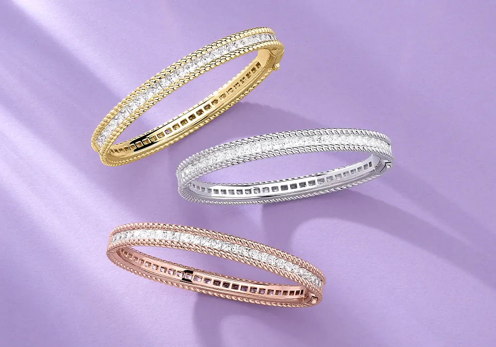 Three APM MONACO Torsade bangles in yellow gold, silver, and rose gold on a light purple background