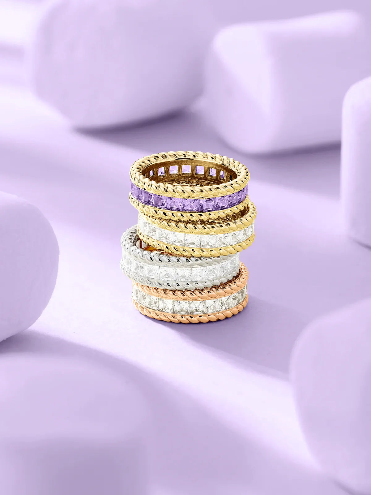 Stack of APM Monaco Torsade gold and rose gold rings with purple gemstone on a light purple background