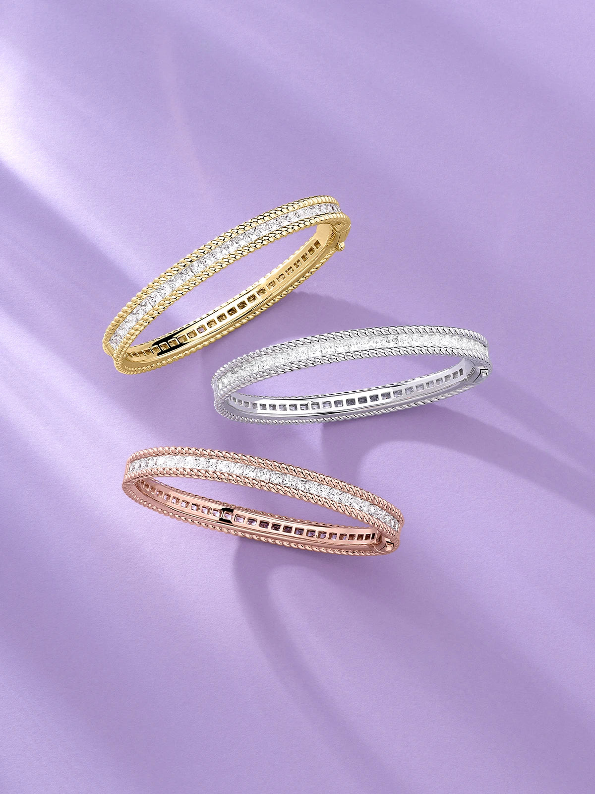 Three APM MONACO Torsade bangles in yellow gold, silver, and rose gold on a light purple background