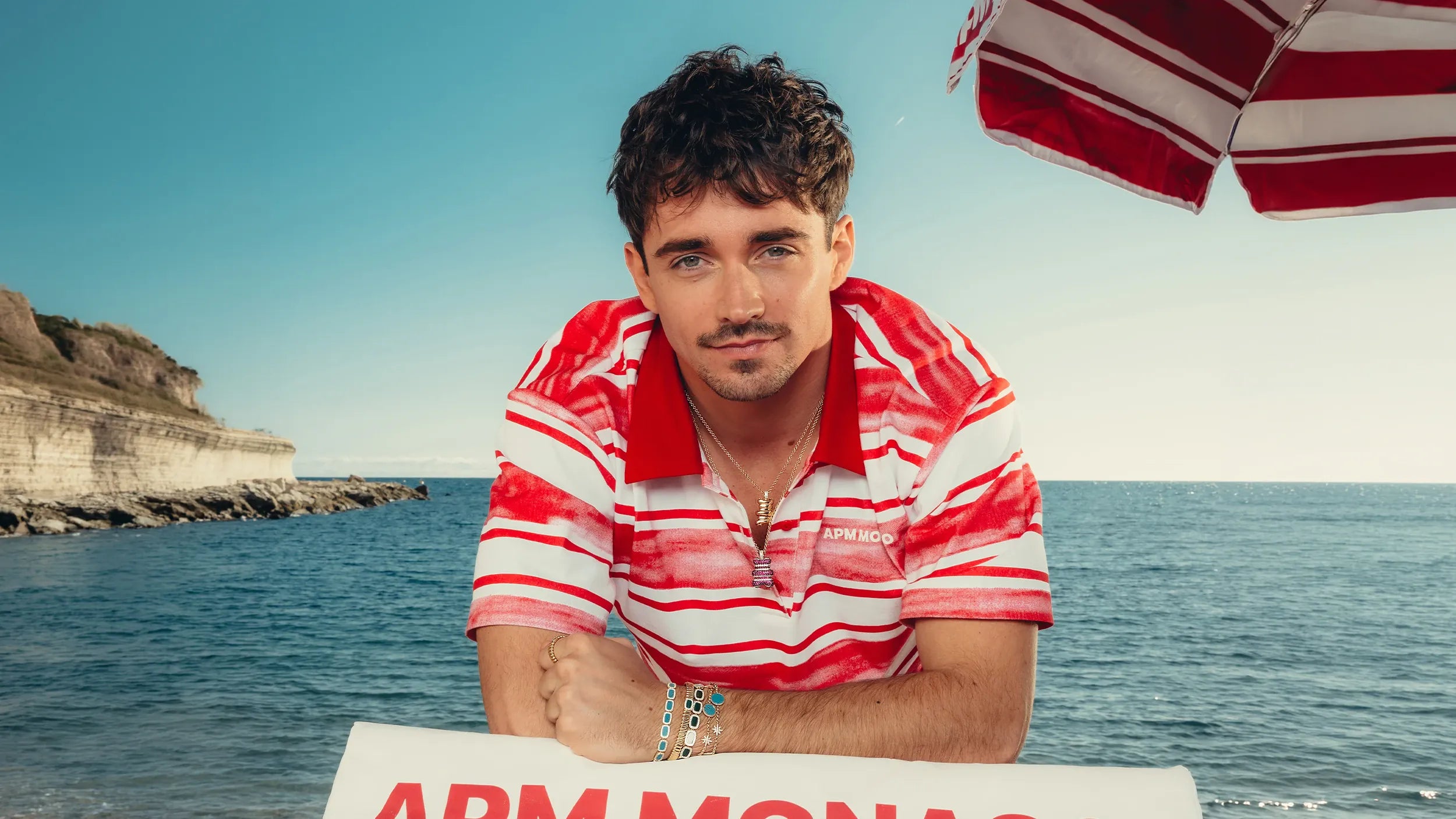 Charles in a red and white striped shirt with APM MONACO jewelry set sitting by the ocean with an umbrella