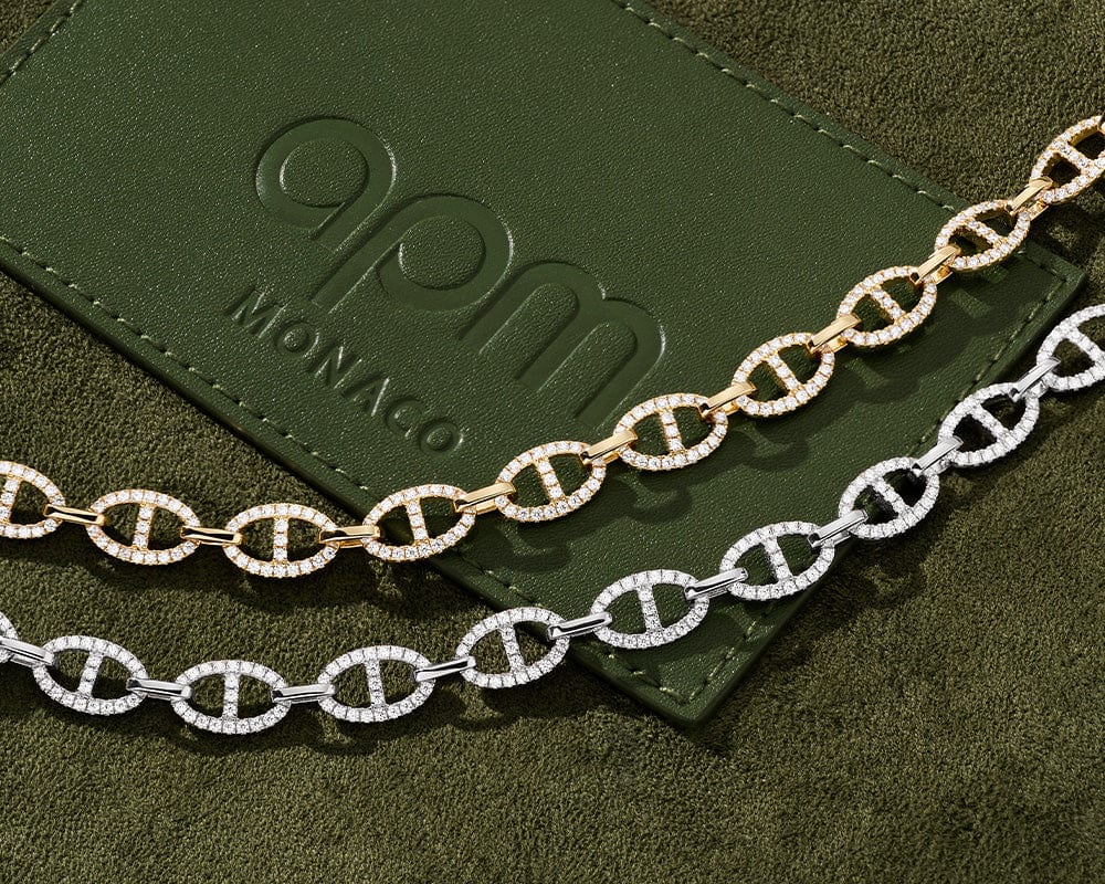 Two chain necklaces, one gold and one silver, on a green surface with APM MONACO branding