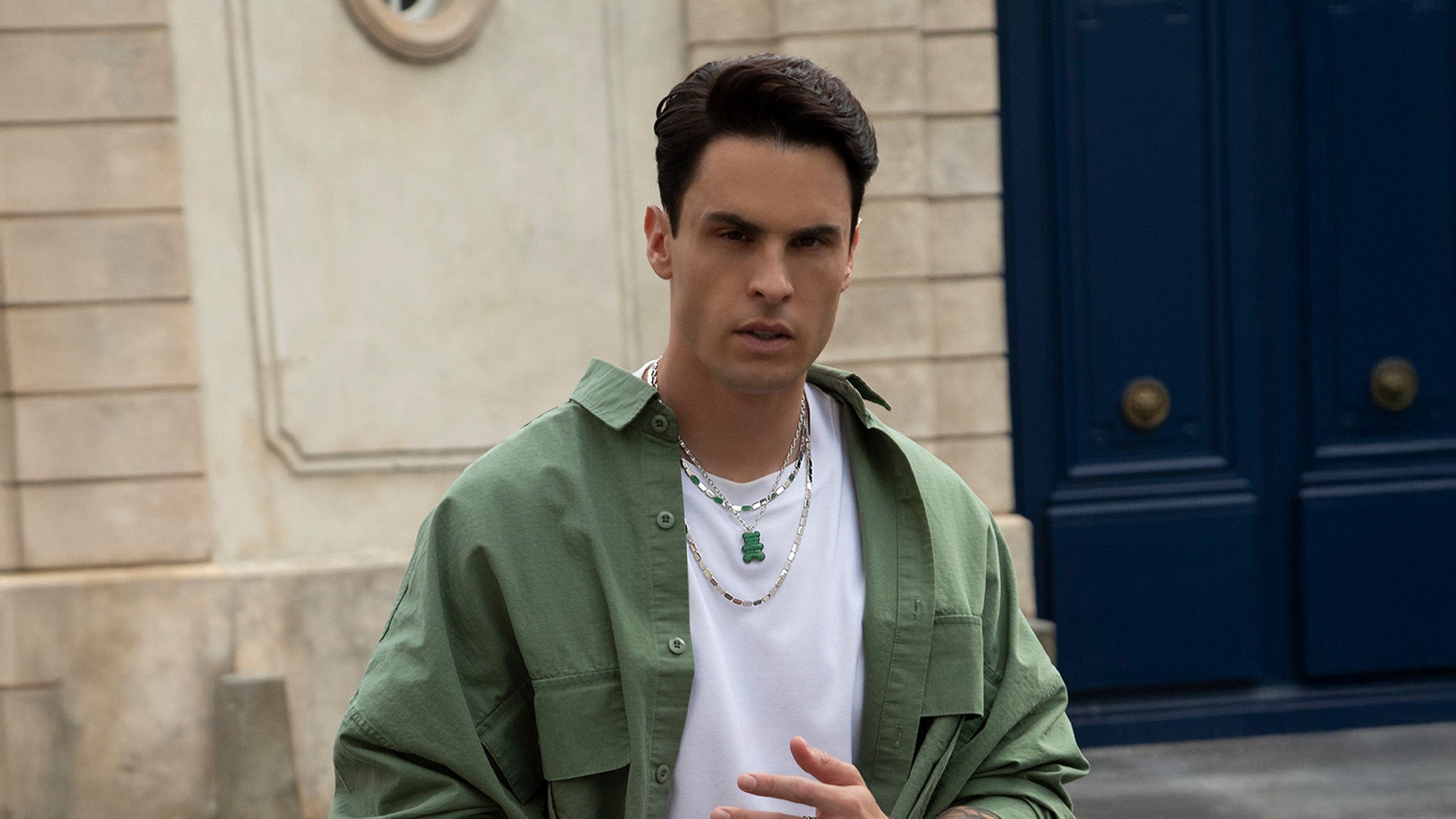 Man wearing yummy and yacht club necklaces standing in front of a blue door.