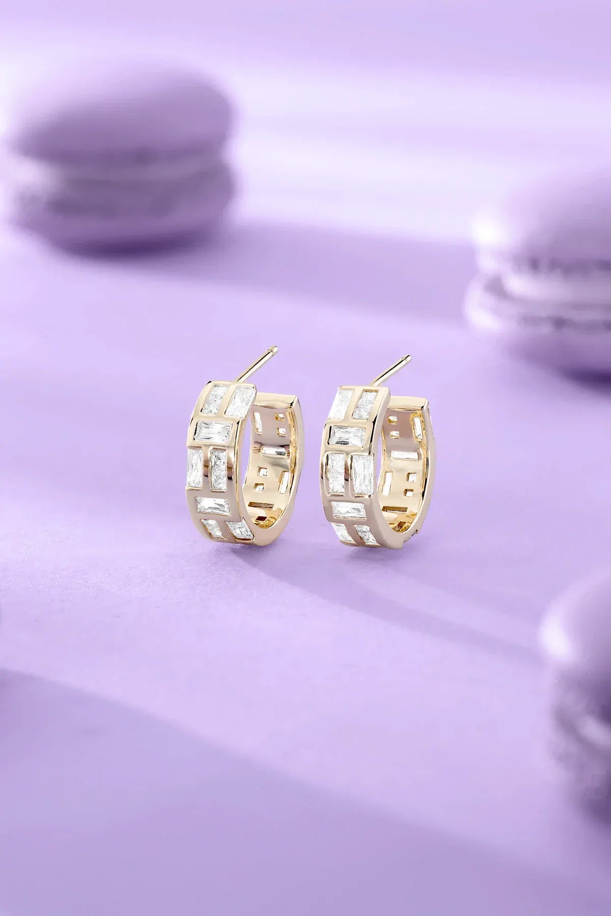 APM MOANCO yellow gold hoop earrings on a light purple surface with blurred macarons in the background